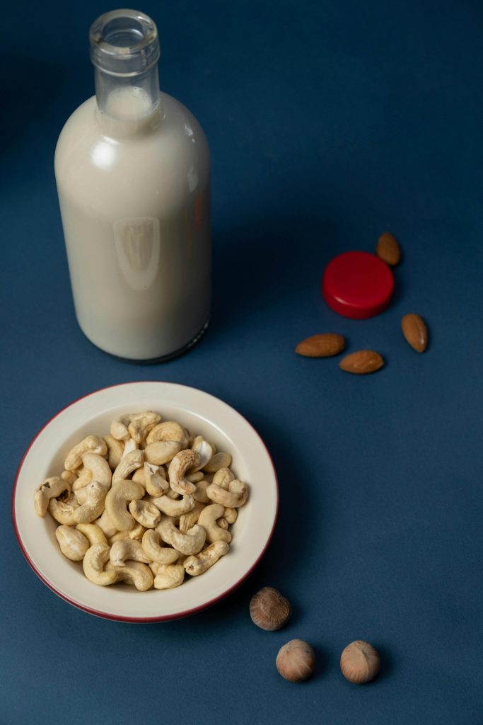 A bottle of vegan cashew milk with a bowl of cashews and mixed nuts on a blue background.